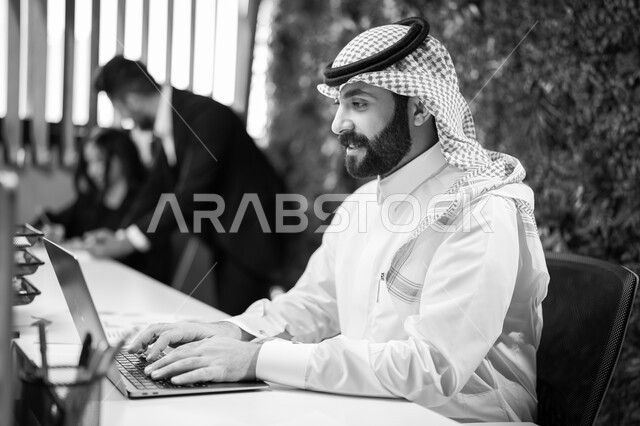 تعابير وايماءات الاندماج والتركيز، صورة بالأسود والأبيض لرجل عربي خليجي سعودي مبتسم يرتدي الشماغ والثوب التقليدي يقوم بالعمل على الحاسب المحمول، إدخال البيانات والمعلومات الكترونيا، شركات ومكاتب سعودية، خلفية المكتب