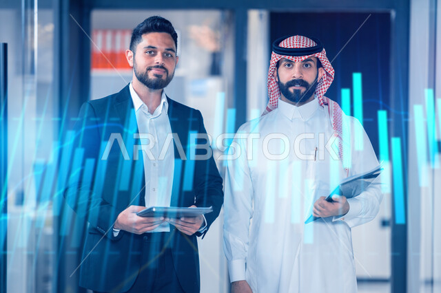 Developing a business strategy and exchanging ideas, standing with gestures of confidence and pleasure to increase the trading of financial profits, monitoring the economy via the holographic chart of the Saudi market, two Saudi Gulf Arab co-workers monitoring the rise and fall of stock exchange stocks via a tablet, company background.