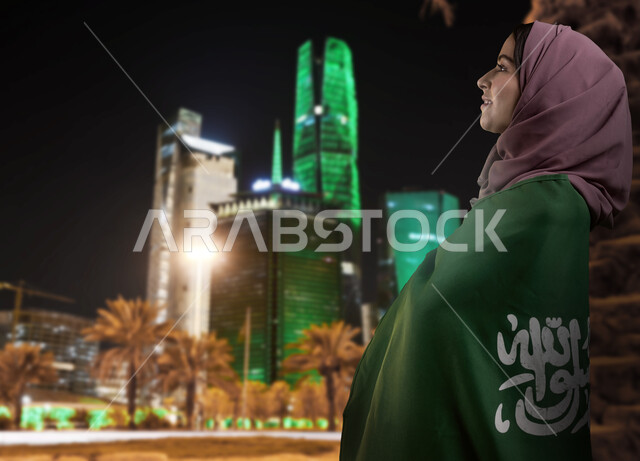 ذكرى العيد الوطني السعودي 23 سبتمبر, الفخر و الاعتزاز بالانتماء للمملكة, صورة من الجانب لامرأة عربية سعودية خليجية ترتدي العباءة والحجاب وتضع علم السعودية على كتفيها للاحتفال بيوم العلم 11مارس, خلفية أبراج مدينة الرياض المضيئة ليلا