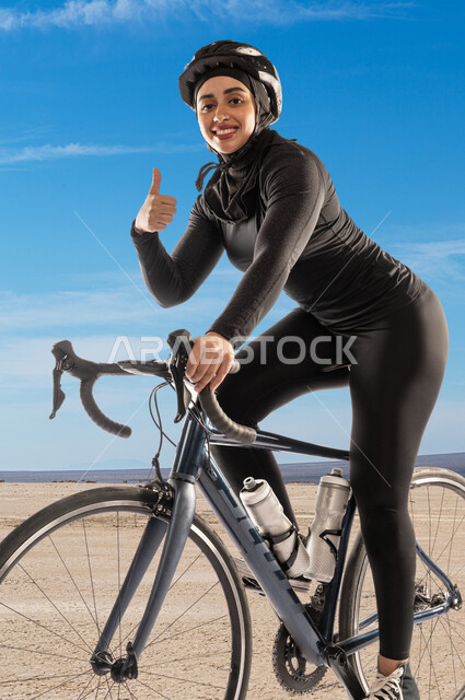 ممارسة رياضة ركوب الدراجة الهوائية، فتاة سعودية عربية خليجية مبتسمة ترتدي الملابس الرياضية وخوذة الحماية ترفع اصبع الابهام للاعلى دليل القبول والرضا، نشاط بدني للمحافظة على اللياقة الجسدية، أنشطة رياضية نسائية في السعودية، جودة الحياة الصحية، خلفية السماء الزرقاء