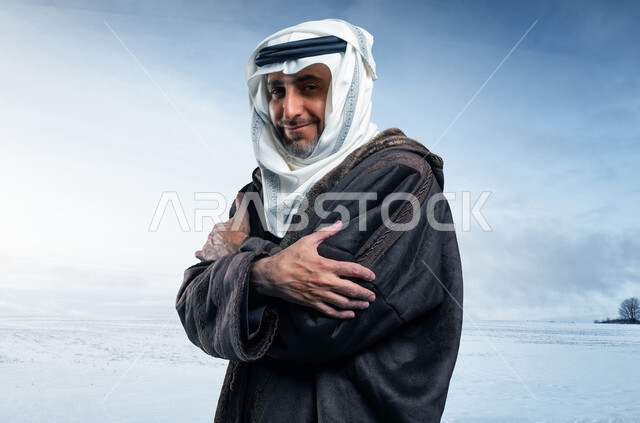إيماءات الشعور بالصقيع والقشعريرة والتجمد، الإحساس الشديد بالبرد في الأجواء الشتوية والثلجية، صورة من الجانب لرجل مسن عربي خليجي سعودي يرتدي الفروة والغترة التقليدية يقف بأيدي متقاطعة في أرض ممتلئةبالثلوج