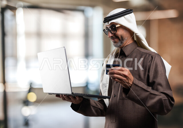 The possibility of paying fees and bills online, using modern technologies and means, an elderly Saudi Arabian Gulf man wearing traditional clothing holding a laptop and bank card in his hand at the workplace, paying via electronic debit cards, lifestyle in Saudi Arabia