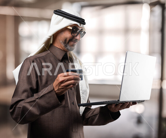 إمكانية تسديد الرسوم والفواتير عبر الانترنت، استخدام التقنيات والوسائل الحديثة، رجل كبير بالسن عربي خليجي سعودي يرتدي الزي التقليدي يمسك بيده اللابتوب والبطاقة البنكية ضمن مقر العمل، الدفع عبر بطاقات الصراف الالكترونية، نمط الحياة في السعودية