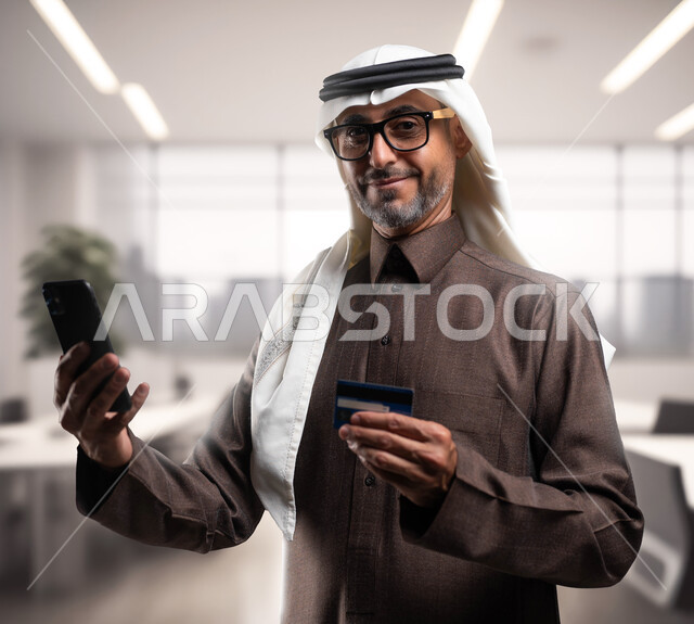 Looking at the camera with happy gestures using modern and advanced means, electronic payment via the Internet, a smiling old Saudi Gulf Arab man wearing the traditional thobe and ghutra holding in his hand a mobile phone and a bank credit card at the workplace.