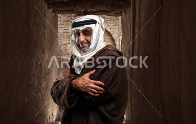 الإحساس الشديد بالبرد في الأجواء الشتوية داخل منزل حجري وأثري قديم، صورة مقربة من الجانب لرجل مسن عربي خليجي سعودي يرتدي الفروة والغترة التقليدية يقف بأيدي متقاطعة، إيماءات الشعور بالصقيع والقشعريرة.