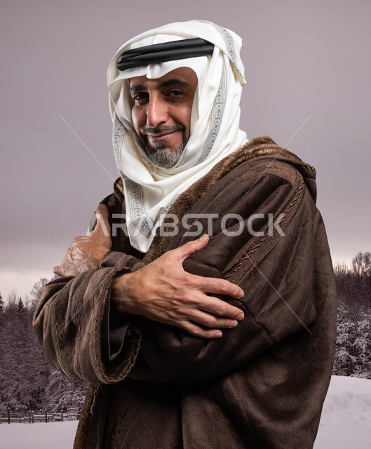 إيماءات الشعور بالصقيع والقشعريرة والتجمد، الإحساس الشديد بالبرد في الأجواء الشتوية والثلجية، صورة من الجانب لرجل مسن عربي خليجي سعودي يرتدي الفروة والغترة التقليدية يقف بأيدي متقاطعة في أرض ممتلئةبالثلوج