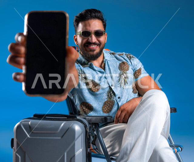 قضاء عطلة صيفية ترفيهيه وممتعة, استخدام الأجهزة والتقنيات الحديثة, عرض خدمات وتطبيقات السياحة والسفر, بورتريه لشاب عربي خليجي سعودي مبتسم يرتدي الزي الكاجوال والنظارة الشمسية يجلس بجانب الحقيبة ويمسك الهاتف المحمول بشاشة سوداء فارغة, خلفية زرقاء