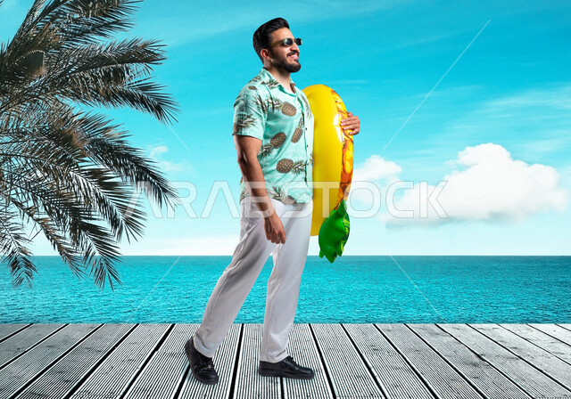 التسلية في إحدى المنتجعات الترفيهيه, أسلوب الحياة في العطلة الصيفية، شاب عربي خليجي سعودي مبتسم يرتدي الزي الكاجوال والنظارة يمسك بيده عوامة السباحة بإيماءات البشاشة والسرور, قضاء أوقات ممتعه في شواطئ السعودية