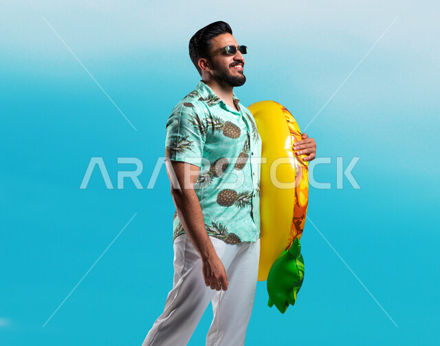 Swimming equipment and tools, spending fun times on the beaches of the summer vacation, portrait of a smiling Saudi Gulf Arab young man wearing casual clothes and glasses holding a float in his hand with gestures of joy and happiness, blue background