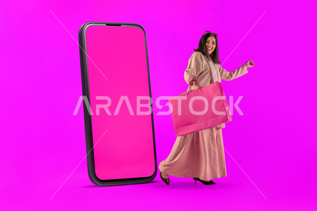 التسوق والتبضع الالكتروني، موك أب لمستطيل وردي كبير، اقتناء المستلزمات والهدايا، بورتريه لإمرأة عربية خليجية سعودية مبتسمة ترتدي عباءة معاصرة تحمل بيدها أكياس ورقية تقف بجانب مجسم لهاتف محمول كبير بشاشة فارغة، مفهوم الموضة والأناقة، خلفية وردية