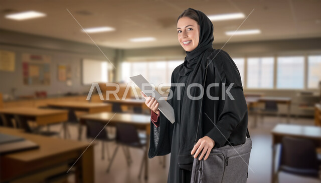 جودة الحياة في المدارس والجامعات السعودية، طالبة جامعية خليجية عربية سعودية مبتسمة ترتدي العباءة السوداء تحمل بيدها الكتب والحقيبة الدراسية، أهمية التعليم في بناء مستقبل متطور للمجتمع السعودي، النظر إلى الكاميرا بإيماءات السرور، خلفية الفصل الدراسي
