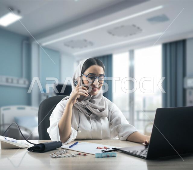 استشارات صحية عبر الجوال، تقديم الخدمات الطبية عن بعد، صورة مقربة لطبيبة عربية خليجية سعودية مبتسمة ترتدي الحجاب والمعطف الابيض والنظارات الطبية تقوم بالعمل على الحاسب المحمول، مفهوم الطب والرعاية الصحية، خلفية ضبابية للعيادة