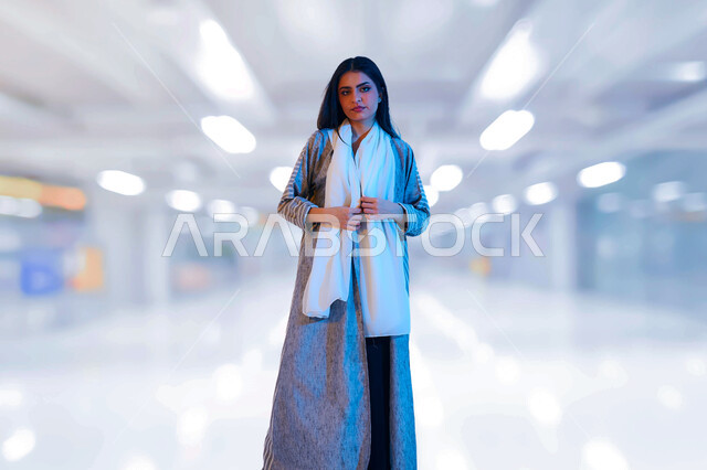 مفهوم الأناقه والأنوثه، إمرأه عربية سعوديه خليجية ترتدي عباءه معاصرة مع شال ابيض تقف بانوثة وتنظر الى الكاميرا، الاستفادة من العروض والتخفيضات التجارية ،الاستمتاع بالشراء والتسوق، قضاء وقت ممتع في التسوق من المول التجاري، خلفية ضبابية لمول