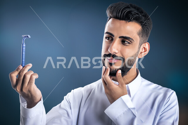 العناية بالبشرة والاهتمام بالمظهر الخارجي، المحافظة على الأناقة والنظافة الشخصية، بورتريه مقرب لرجل عربي خليجي سعودي يرتدي الثوب التقليدي يمسك بيده شفرة الحلاقة الرجالية وينظر إليها بإيماءات الإعجاب والسرور، خلفية سوداء