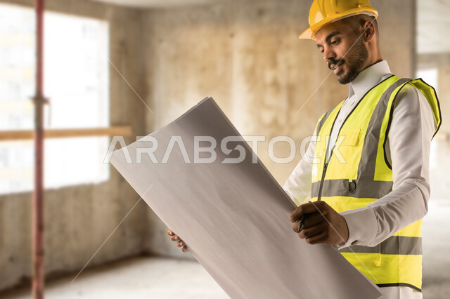 الاشراف على تطبيق الخطط والمشاريع الهندسيه المعمارية في السعودية، مهندس عربي خليجي سعودي مبتسم يرتدي الخوذة وسترة الحماية يقوم بمشاهدة مخطط البناء في مقر عمله، العمل في القطاع الهندسي، مفهوم الهندسة والانشاءات، خلفية مبنى قيد الانشاء