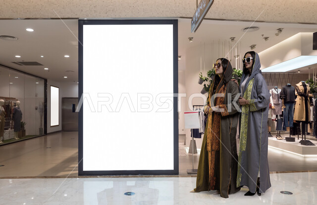 موك أب لمستطيل أبيض كبير، أزياء وإكسسوارات نسائيه، شابتان عربيتان سعوديتان خليجيتان، ترتديان العباءة الخليجية العصرية والنظارات الشمسية، السعاده والمتعة بالتسوق مع الاصدقاء، مفهوم الموضة والأناقة، خلفية المحلات التجارية