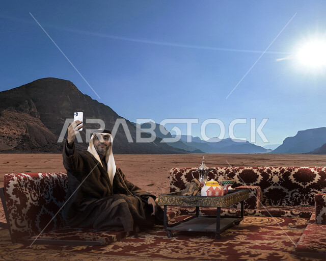 جلسات البر الشعبية البدوية ،أجواء التخييم الهادئة في فصل الشتاء، توثيق اللحظات السعيدة بالتقاط صور تذكارية عبر الهاتف المحمول، رجل كبير السن عربي سعودي خليجي يرتدي اللباس الشتوي الدافئ والزي التقليدي يحمل بيده الجوال في النهار، خلفية الصحراء والمرتفعات الجبلية