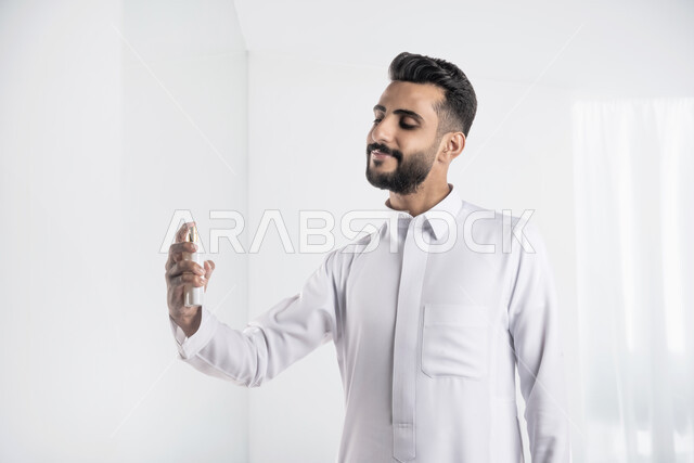 عطورات خليجية سعودية, الاستعداد للذهاب والخروج من المنزل, مفهوم التعطر والتطيب, استخدام الزيوت العطرية الطبيعية, بورتريه مقرب لرجل عربي خليجي سعودي مبتسم يرتدي الثوب التقليدي يقوم برش العطر, خلفية بيضاء