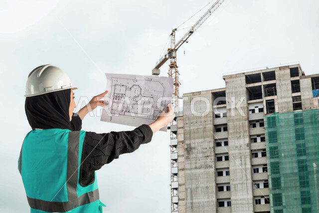 مشاركة النساء السعوديات في مجال الهندسات، متابعة المشروع وتدقيق المخططات، صورة من الجانب لمهندسة معمارية عربية خليجية سعودية ترتدي الخوذة وسترة الوقاية تحمل بيده مخطط البناء في مقر العمل، الاشراف والرقابة على الانشاءات، التحقق من تطبيق المشاريع والخطط الهندسيه المعمارية بشكل صحيح