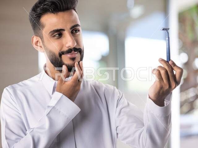 المحافظة على الأناقة والنظافة الشخصية، العناية بالبشرة والاهتمام بالمظهر الخارجي، بورتريه مقرب لرجل عربي خليجي سعودي يرتدي الثوب التقليدي يمسك بيده شفرة الحلاقة الرجالية وينظر إليها بإيماءات الإعجاب والسرور