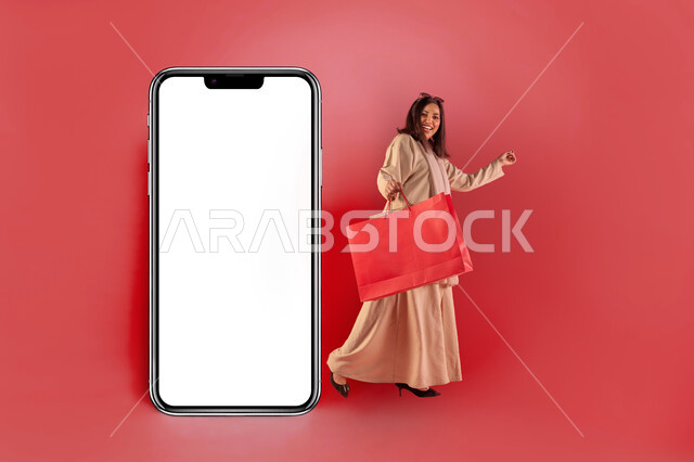التسوق والتبضع الالكتروني، موك أب لمستطيل أبيض كبير، اقتناء المستلزمات والهدايا، بورتريه لإمرأة عربية خليجية سعودية مبتسمة ترتدي عباءة معاصرة تحمل بيدها كيس ورقي أحمر تقف بجانب مجسم لهاتف محمول كبير بشاشة بيضاء فارغة، مفهوم الموضة والأناقة، خلفية حمراء