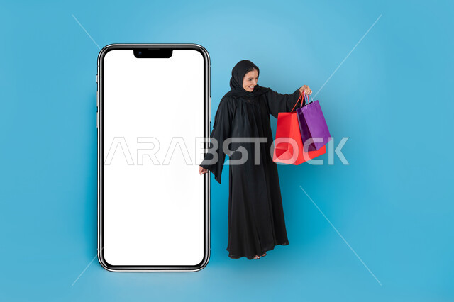 موك أب لمستطيل أبيض كبير، اقتناء المستلزمات والهدايا، بورتريه لإمرأة عربية خليجية سعودية محجبة ترتدي عباءة سوداء عصرية تحمل بيدها الأكياس الملونة تقف بجانب مجسم لهاتف محمول كبير بشاشة فارغة، مفهوم الموضة والأناقة، التسوق والتبضع الالكتروني، خلفية زرقاء