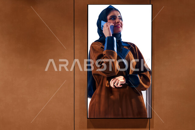 انعكاس على المرآه  لصورة امرأه عربيه خليجية سعودية محجبة تقوم بالتحدث على الهاتف، دردشة ومحادثات مع الأهل والأصدقاء، استخدام الهواتف الذكية والتقنيات الحديثة، خلفية باللون البني