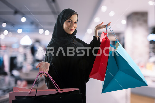 التبضع وشراء المستلزمات، الاستفادة من العروض الموسمية والتخفيضات, صورة مقربة لامرأة عربيه سعودية خليجية محجبة ترتدي عباءه سوداء تحمل بيدها أكياس التسوق الملونة، زيارة المولات التجارية، جودة الحياة في السعودية
