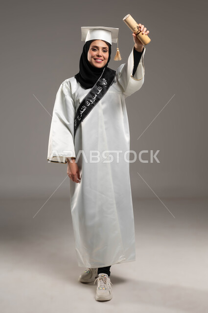 Fulfilling the dream of graduating from university with gestures of joy and pleasure, a close-up portrait of a Saudi Gulf Arab student wearing a graduation uniform and hat, raising her hands enthusiastically and holding the university document, full-length body portrait, gray background