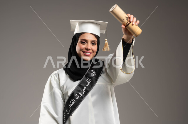 الاحتفال بتحقيق حلم التخرج من الجامعة بايماءات الفرح والسرور, بورتريه مقرب لطالبة عربية خليجية سعودية ترتدي زي التخرج والقبعة ترفع يديها بحماس وتحمل الوثيقة الجامعية, خلفية رمادية