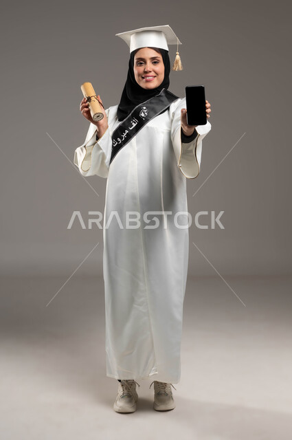 استخدام الأجهزة والتقنيات الحديثة, أهمية الدراسة الجامعية, بورتريه لشابة عربية سعودية خليجية ترتدي القبعة وعباءة التخرج باللون الأبيض بايماءات السعادة تمسك بيدها شهادة التخرج, عرض الهاتف المحمول بشاشة سوداء فارغة, صورة الجسم بالطول الكامل, خلفية رمادية
