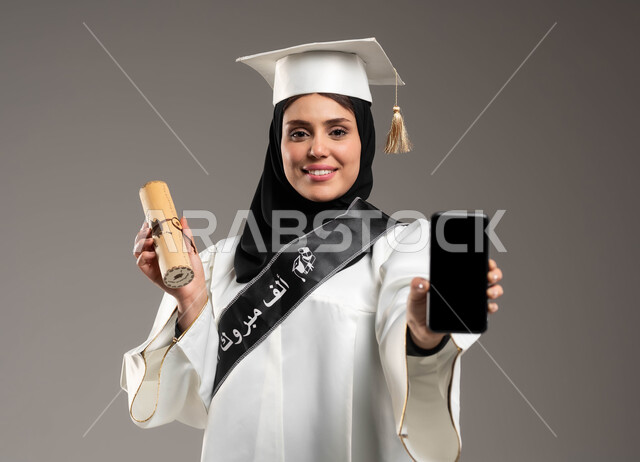 أهمية الدراسة الجامعية, استخدام الأجهزة والتقنيات الحديثة, بورتريه لخريجة جامعية عربية سعودية خليجية ترتدي القبعة وعباءة التخرج باللون الأبيض بايماءات السعادة تمسك بيدها شهادة التخرج, عرض الهاتف المحمول بشاشة سوداء فارغة, خلفية رمادية