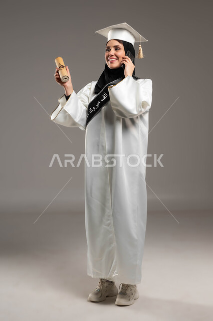 استخدام الأجهزة والتقنيات الحديثة, الاحتفال بالتخرج من الجامعة, إجراء مكالمة هاتفية عبر الجوال, بورتريه لخريجة جامعية عربية سعودية خليجية ترتدي العباءة وقبعة التخرج باللون الأبيض بإيماءات الفرح والسعادة وتمسك بيدها شهادة النجاح والتفوق, صورة الجسم بالطول الكامل, خلفية رمادية