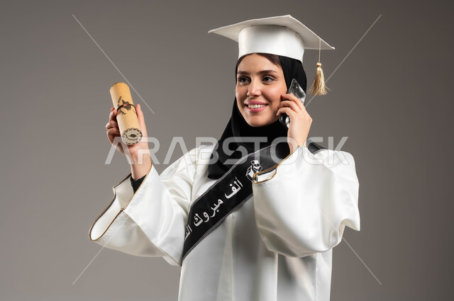 الاحتفال بالتخرج من الجامعة, إجراء مكالمة هاتفية عبر الجوال, بورتريه مقرب لخريجة جامعية عربية سعودية خليجية ترتدي العباءة وقبعة التخرج باللون الأبيض, ايماءات الفرح والسعادة, تمسك بيدها شهادة النجاح والتفوق, استخدام الأجهزة والتقنيات الحديثة, خلفية رمادية