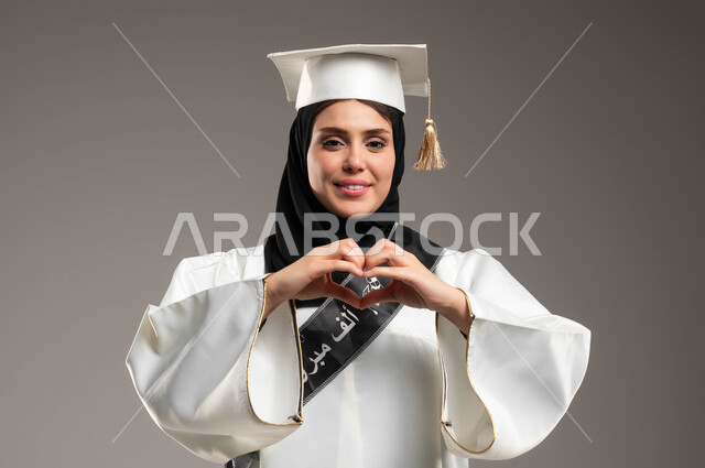 The importance of university studies and the joy of achieving success, university events and celebrations, a close-up portrait of a Saudi Arabian Gulf student wearing a white graduation gown and hat, raising her hands in the shape of a heart with gestures of love and contentment, education in the Kingdom of Saudi Arabia, gray background