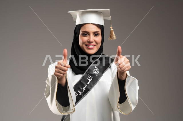 احتفالات التخرج الجامعية, التعليم في المملكة العربية السعودية, بورتريه مقرب لطالبة عربية سعودية خليجية ترفع يديها ترتدي ثوب التخرج الابيض والقبعة بإيماءات الجودة والامتياز والرضا, النظر إلى الكاميرا بسعادة وفرح بتحقيق حلم النجاح, خلفية رمادية