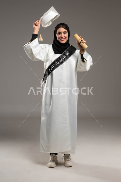 University events and celebrations, gestures of pride and joy in achieving the dream of success, a close-up portrait of a Saudi Arabian Gulf student wearing a white graduation gown, raising her hand up enthusiastically and holding the hat and graduation certificate, full-length body portrait, gray background