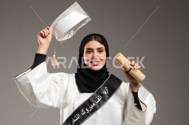 ايماءات الفخر والسرور بتحقيق حلم النجاح, بورتريه مقرب لطالبة عربية سعودية خليجية ترتدي ثوب التخرج الابيض ترفع يدها للأعلى بحماس وتمسك القبعة ووثيقة التخرج, المناسبات والاحتفالات الجامعية, خلفية رمادية