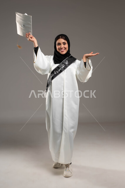 Gestures of pride and pleasure in achieving the dream of success, gestures of joy and happiness at graduation, portrait of a Saudi Arabian Gulf student wearing a white graduation gown and hat, raising her hands up enthusiastically, university events and celebrations, full-length body portrait, gray background