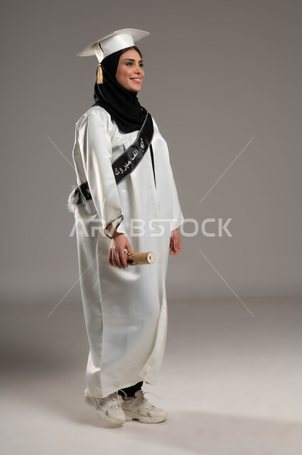 Standing proudly and looking at something, gestures of joy and happiness in reaching the dream of success, portrait of a Saudi Gulf Arab student wearing a white dress and hat holding a graduation document in her hand, university events and celebrations, full-length body portrait, gray background