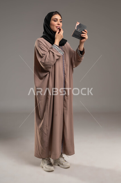 مستحضرات تجميلية نسائية, مكياج تتوريال, الشغف بوضع الميك اب, التسويق لمنتجات التجميل, بورتريه لامرأة عربية خليجية سعودية مبتسمة ترتدي العباءة البنية والحجاب تضع احمر الشفاه وتنظر الى المرآة, صورة الجسم بالطول الكامل, خلفية رمادية