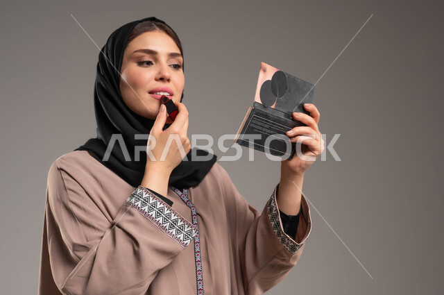 مكياج تتوريال, الشغف بوضع الميك اب, مستحضرات تجميلية نسائية, التسويق لمنتجات التجميل, بورتريه مقرب لامرأة عربية خليجية سعودية مبتسمة ترتدي العباءة البنية والحجاب تضع احمر الشفاه وتنظر الى المرآة, خلفية رمادية