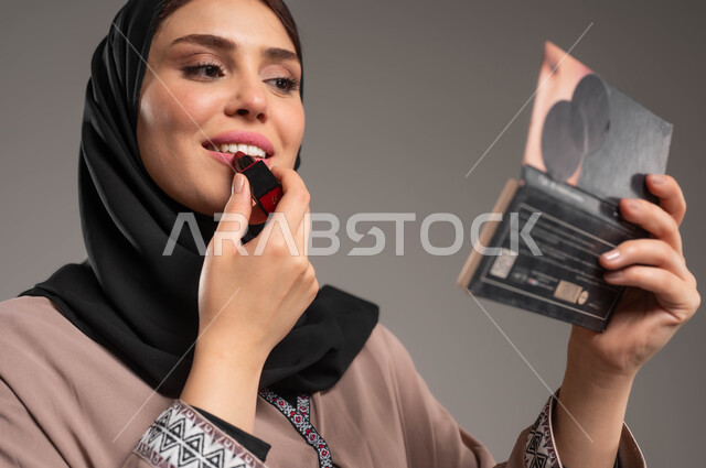 الشغف بوضع الميك اب, مستحضرات تجميلية نسائية, التسويق لمنتجات التجميل, بورتريه مقرب لامرأة عربية خليجية سعودية مبتسمة تضع احمر الشفاه وتنظر الى المرآة ترتدي عبائة بنية, مكياج تتوريال, خلفية رمادية