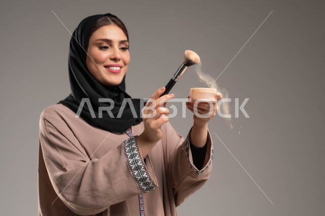 الوقوف بسرور وببشاشة والاستمتاع بالاعتناء بالنفس, مفهوم الاهتمام بالجمال, التسويق لمستحضرات التجميل, بورتريه مقرب لامرأة عربية سعودية خليجية مبتسمة ترتدي العباءة البنية والحجاب تمسك بيدها فرشاة المكياج, خلفية رمادية