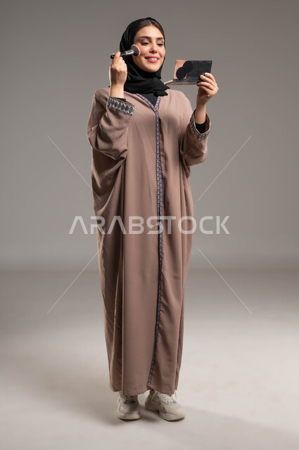 التسويق لمنتجات التجميل, مستحضرات تجميلية نسائية, بورتريه لامرأة عربية خليجية سعودية مبتسمة ترتدي العباءة البنية والحجاب تمسك بيدها إحدى أدوات التجميل(الفرشاة), ميك اب تتوريال, صورة الجسم بالطول الكامل, خلفية رمادية