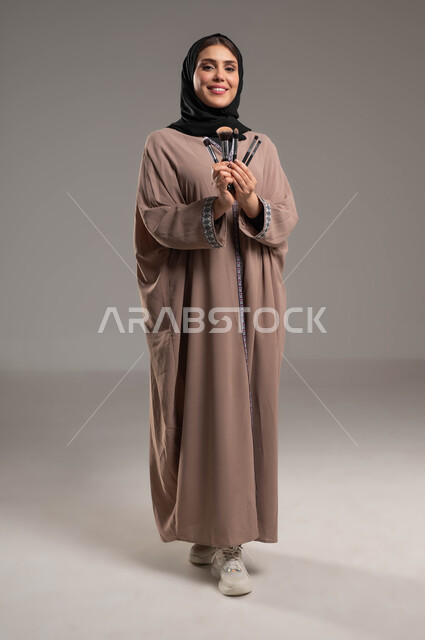 أدوات مكياج تتوريال, مفهوم الاهتمام بالجمال والبشرة والمظهر الخارجي, جودة المنتجات السعودية, بورتريه لامرأة عربية سعودية خليجية مبتسمة ترتدي العباءة البنية والحجاب تمسك بيدها أدوات التجميل, صورة الجسم بالطول الكامل, خلفية رمادية
