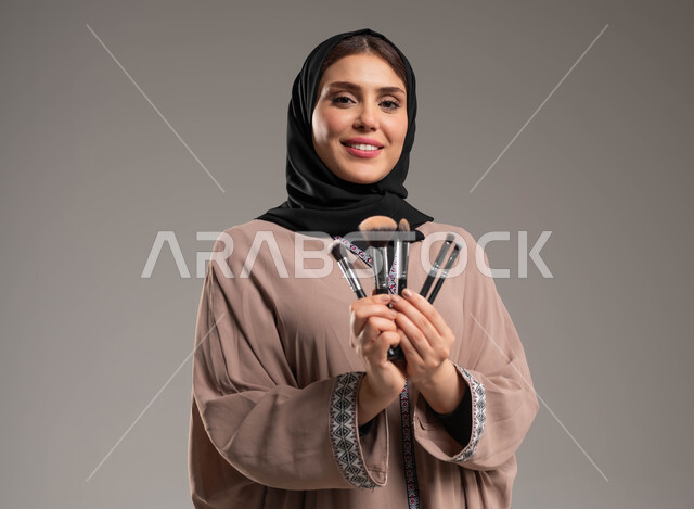 مفهوم الاهتمام بالجمال والبشرة والمظهر الخارجي, جودة المنتجات السعودية, أدوات مكياج تتوريال, بورتريه مقرب لامرأة عربية سعودية خليجية  مبتسمة ترتدي عباءة بنية تمسك بيدها  أدوات التجميل, خلفية رمادية