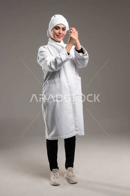 الاهتمام بالمظهر الخارجي، مفهوم العناية  بالبشرة، بورتريه لامرأة عربية خليجية سعودية ترتدي روب استحمام أبيض و منشفة بيضاء تضع كريم الترطيب على يديها بإيماءات السرور، استخدام كريمات ومكونات تجميلية، صورة الجسم بالكامل، خلفية رمادية