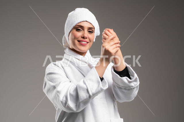 استخدام كريمات و مكونات تجميلية، مفهوم العناية بالبشرة، الاهتمام بالمظهر الخارجي، بورتريه لامرأة عربية خليجية سعودية ترتدي روب استحمام أبيض و منشفة بيضاء تحمل بيدها علبة كريم الترطيب، صورة الجسم بالكامل، خلفية رمادية