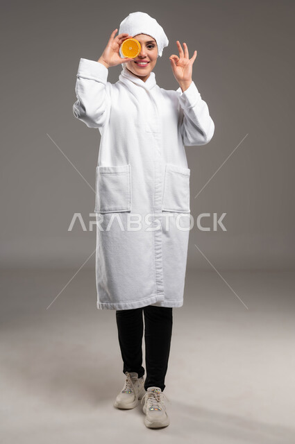 النظر للكاميرا بايماءات السرور، حمية غذائية صحية للحفاظ على الرشاقة،  اتباع نظام غذائي صحي، بورتريه لمرأة عربية خليجية سعودية مبتسمة ترتدي روب استحمام ومنشفة بيضاء تحمل شريحة برتقال، فيتامينات مغذية للجسم، فطور صباحي طبيعي مفيد، صورة الجسم بالكامل، خلفية رمادية